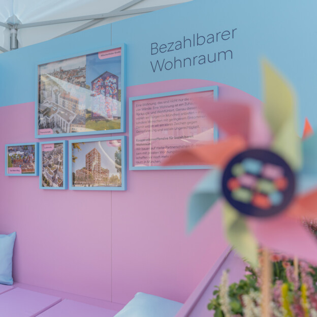 Blick auf einen Messestand auf dessen Rückwand steht "Bezahlbarer Wohnraum". Daneben hängen Fotografien von Neubauten und Quarteieren. IM Vordergrund sieht man ein Windrad mit dem Münchner Wohnen Logo.