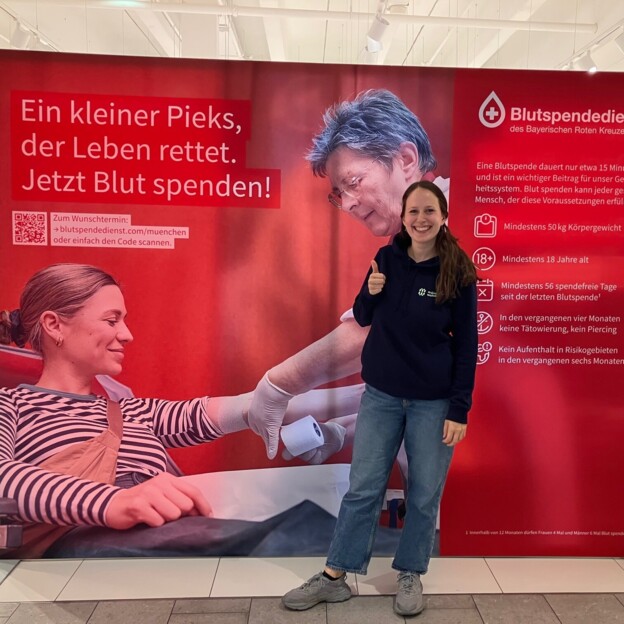 Eine Frau im Pullover der Münchner Wohnen steht vor einem Plakat eines Blutspendedienstes auf dem steht "Ein kleiner Pieks, der Leben rettet. Jetzt Blut spenden!".