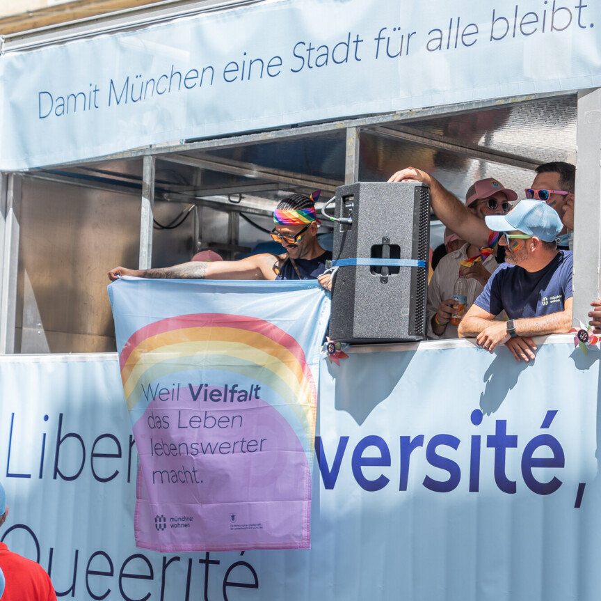 Blick auf einen Truck der im Münchner Wohnen Design geschmückt ist. Auf dem Truck stehen feiernde Menschen, einer von Ihnen hält eine Fahne auf der steht "Weil Vielfalt das Leben lebenswerter macht.".