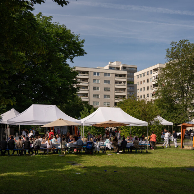 Blick auf einen großen Innenhof mit vielen Bäumen und Wiese. Dort stehen mehrere überdachte Bänke und Tische an den Nachbar*innen gemeinsam feiern.