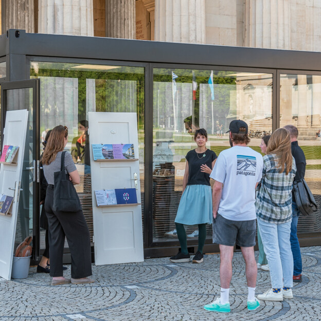 Eine Menschengruppe steht vor einem gläsernen Container, der Dachziegel enthält Eine Frau erklärt der Gruppe das Konzept der Münchner Wohnen.