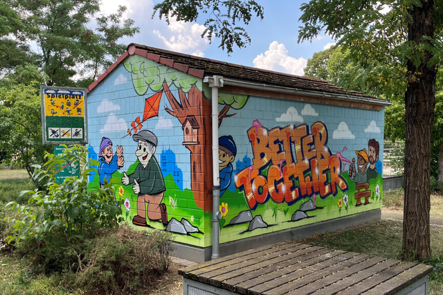 Ein Stromhäuschen steht auf einer Grünfläche. Die Wände sind bunt bemalt und zeigen Menschen, die zusammen spielen und sich unterhalten. Auf einer Wand steht "Better together".
