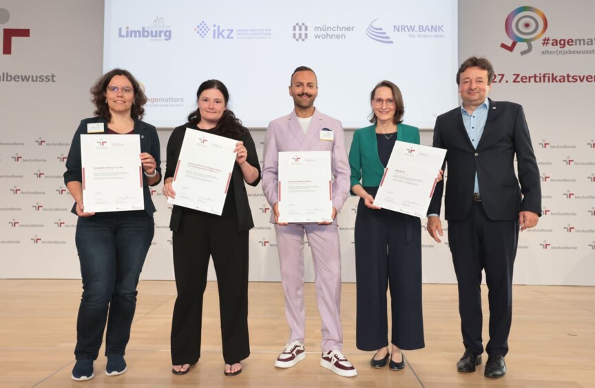 Fünf Unternehmensvertreter*innen stehen auf einer Bühne und halten feierlich ihre Zertifikatsurkunden in der Hand.