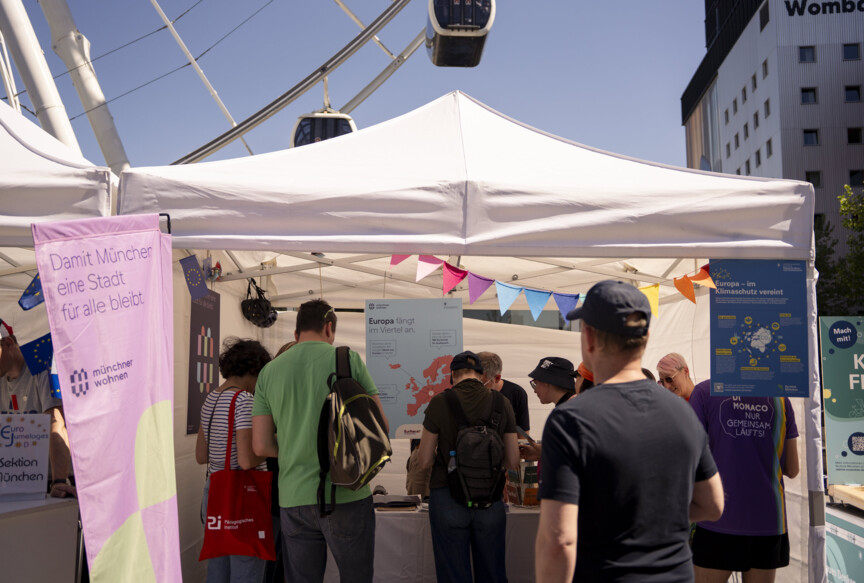 Blick auf den Stand der Münchner Wohnen. Davor sind einige Menschen. Im Hintergrund sieht man die Gondeln des Riesenrads.