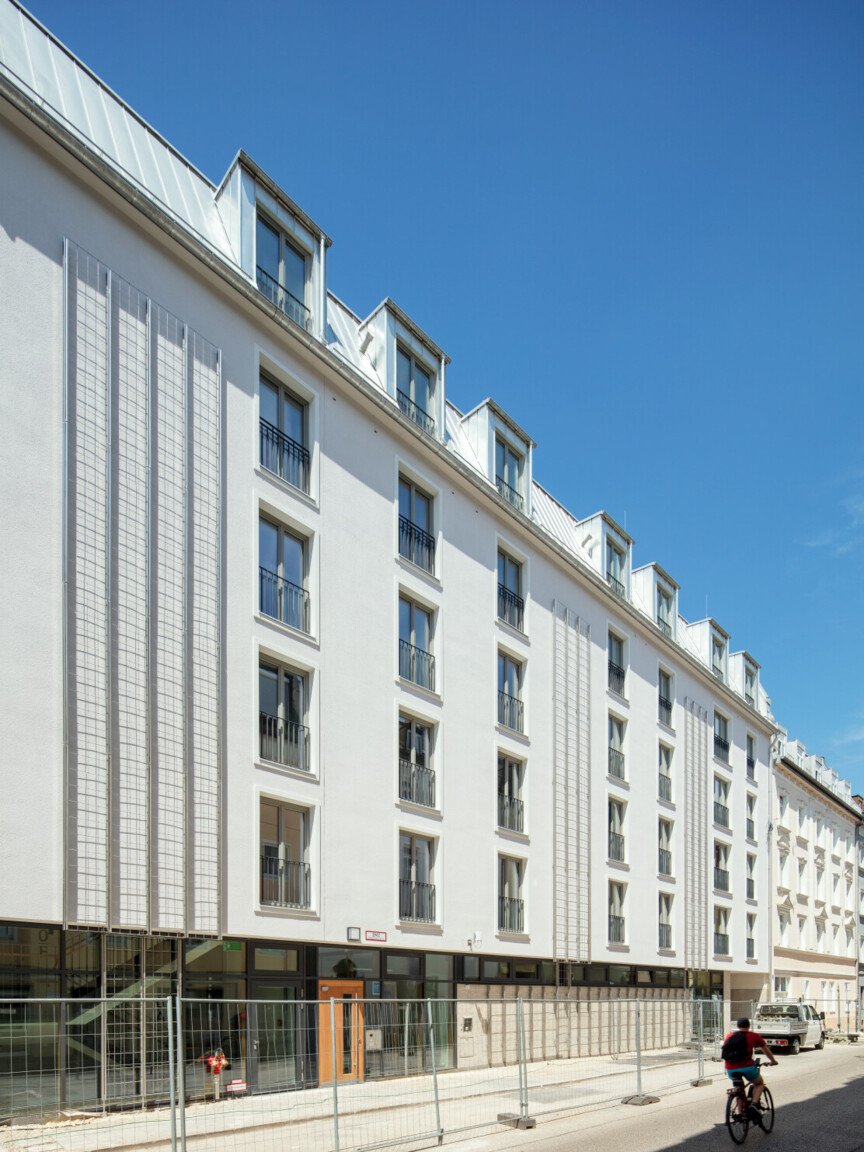 Weiße Hausfassade. Vor dem Erdgeschoss ist ein Bauzaum. Auf der Straße fährt eine Person mit dem Fahrrad vorbei.