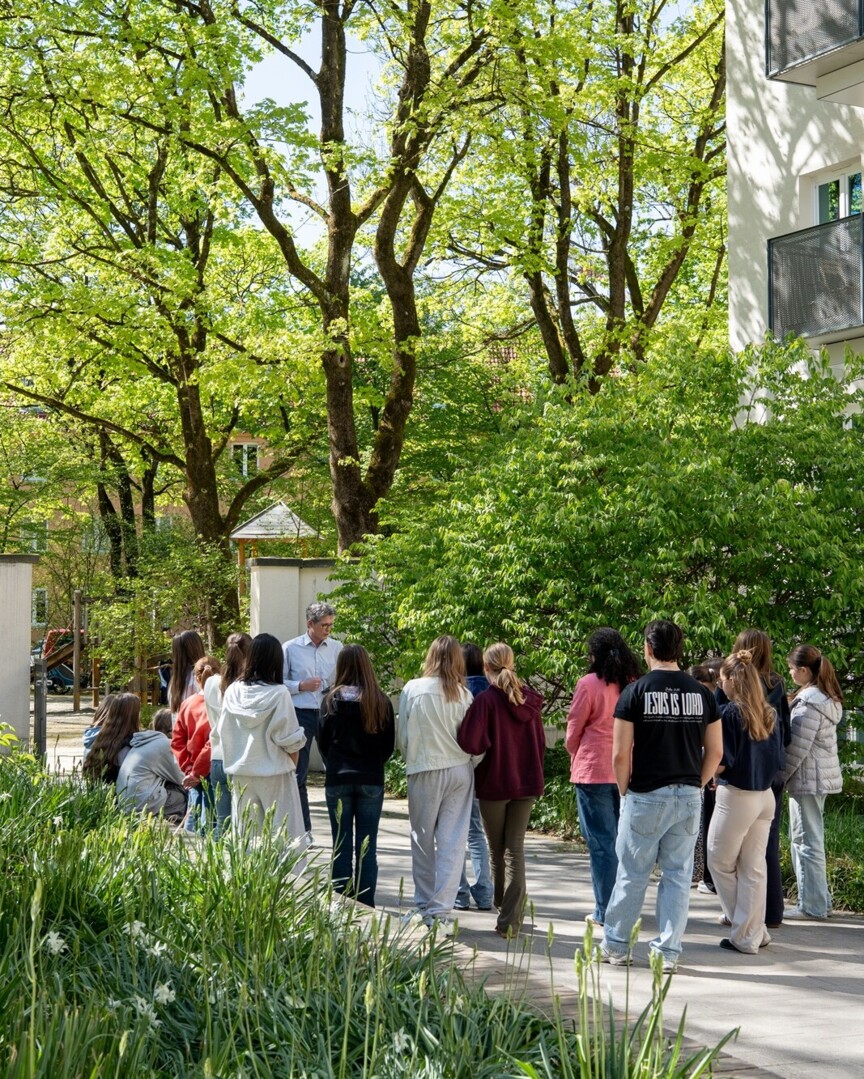 Blick auf eine Menschengruppe die in einer Wohnanlage steht. Ein Mann erklärt etwas.