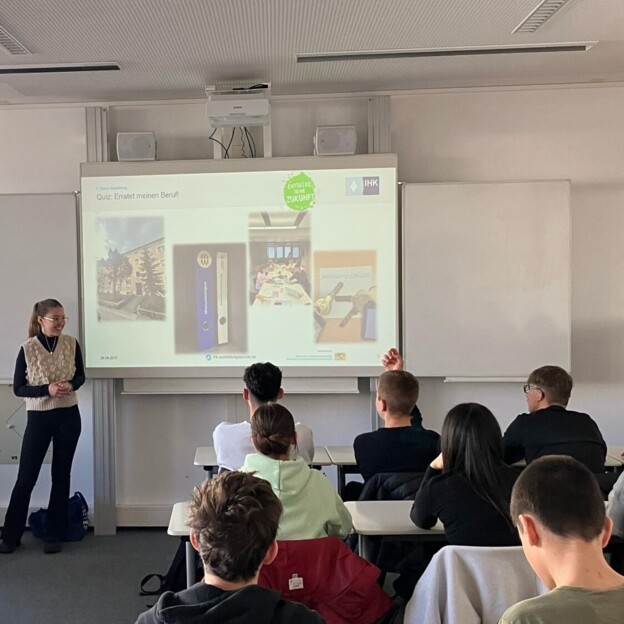 Blick über das Publikum hinweg auf eine Leinwand auf der Impressionen aus dem Arbeitsalltag bei der Münchner Wohnen gezeigt werden. Links daneben steht die Referentin.