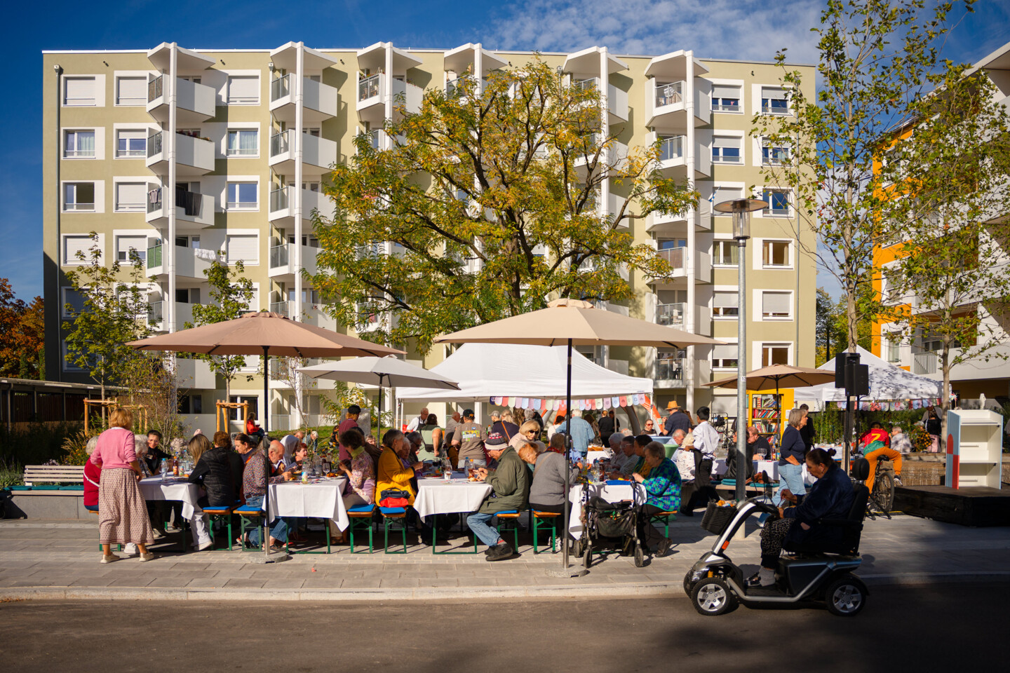Im Vordergrund sitzen viele Menschen auf Bierbänken und unterhalten sich. Im Hintergrund sieht man das modernisierte Mehrfamilienhaus.