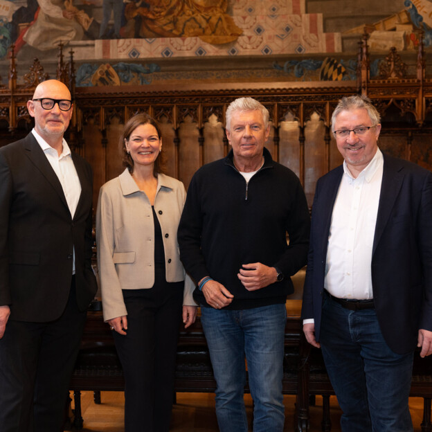 Drei Männer und eine Frau stehen im historischen Rathaussaal Münchens und blicken gemeinsam in die Kamera.