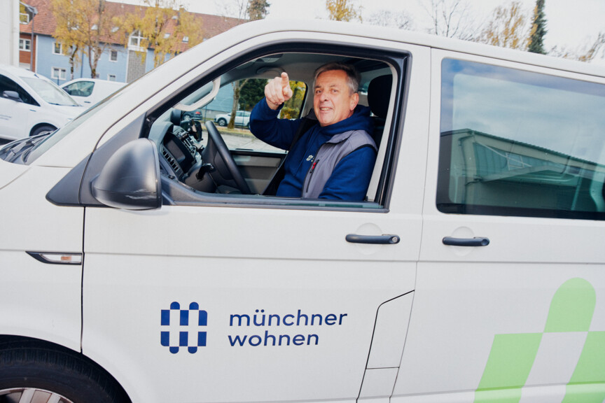 Ein Mann in Arbeitskleidung sitzt in einem weißen Auto mit Münchner Wohnen Logo.