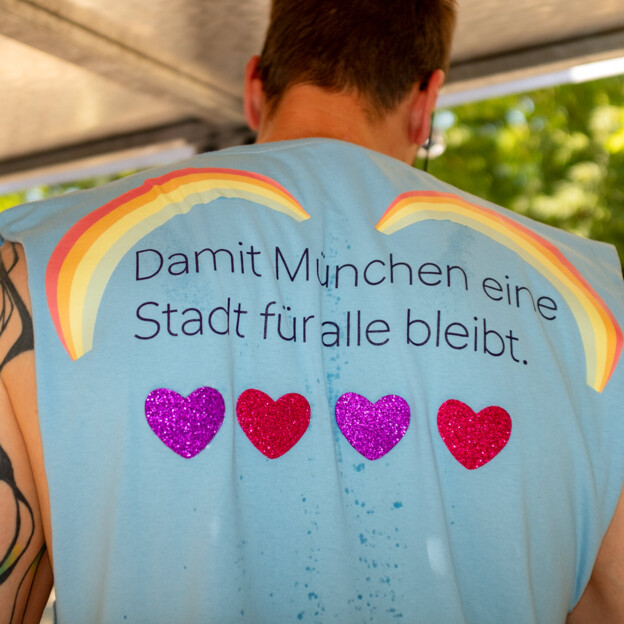 Blick auf den Rücken eines Mannes. Auf seinem bunt geschmückten Shirt steht "Damit München eine Stadt für alle bleibt.".