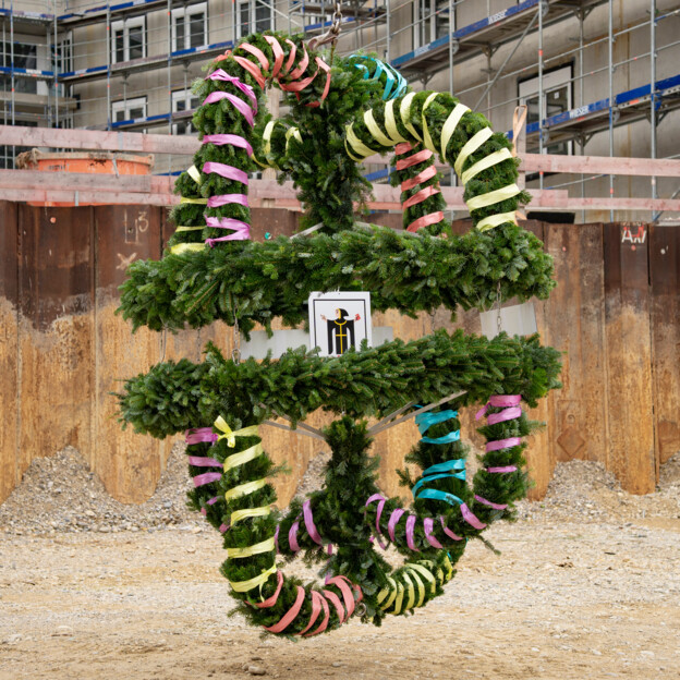 Richtkrone die mit bunten Bändern geschmückt ist. In der Mitte hängt das Münchner Kindl, Münchens Stadtwappen. Im Hintergrund sieht man eine Baustelle.