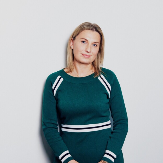 Eine Frau in einem grünen Strickkleid und mit blonden Haaren steht vor einer Wand und lächelt in die Kamera.