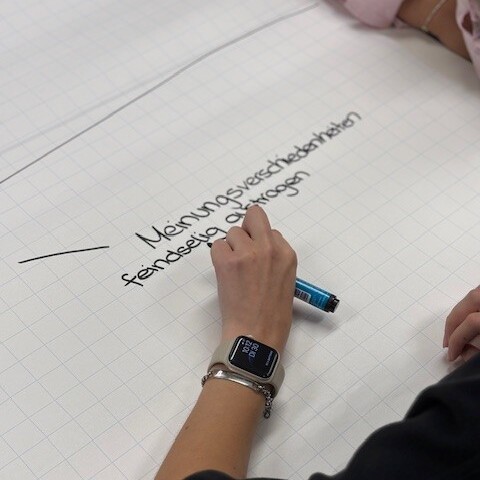 Blick über die Schulter einer Person auf ein großes weißes Blatt Papier auf dem "Meinungsverschiedenheiten feindselig austragen" steht. Die Person hält einen Stift in der Hand.