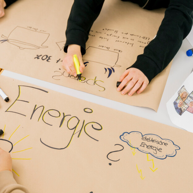 Blick von oben auf zwei Plakate. Hände schreiben die Wörter "Energie" und "sparen". Daneben sind einige Zeichnungen zu sehen.