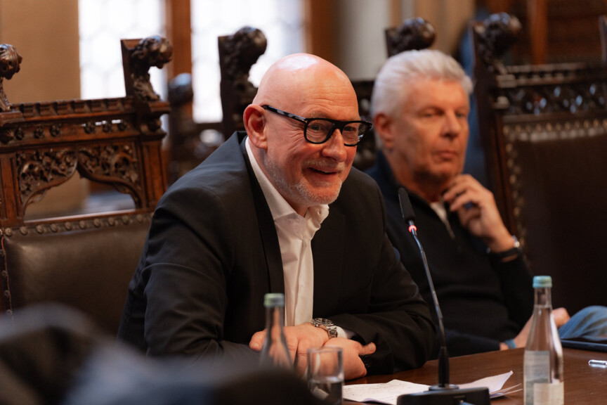 Zwei Männer sitzen nebeneinander an einem Tisch. Der eine beugt sich vor und spricht in ein Tischmikrofon.