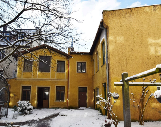 Ansicht des denkmalgeschützten Hauses, dass im Innenhof einer Wohnanlage steht. Das L-förmige Gebäudeist zweistöckig und gelb gestrichen. Die Farbe ist sieht schon älter aus und man erkennt, dass das Gebäude schon älteren Baujahres ist.