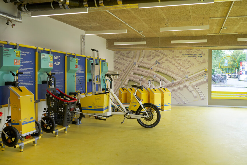 Innenansicht einer Mobilitätsstation. In einem hellen, großen Raum stehen Handkarren und ein Fahrrad. An der Wand befinden sich Informationsplakate. 