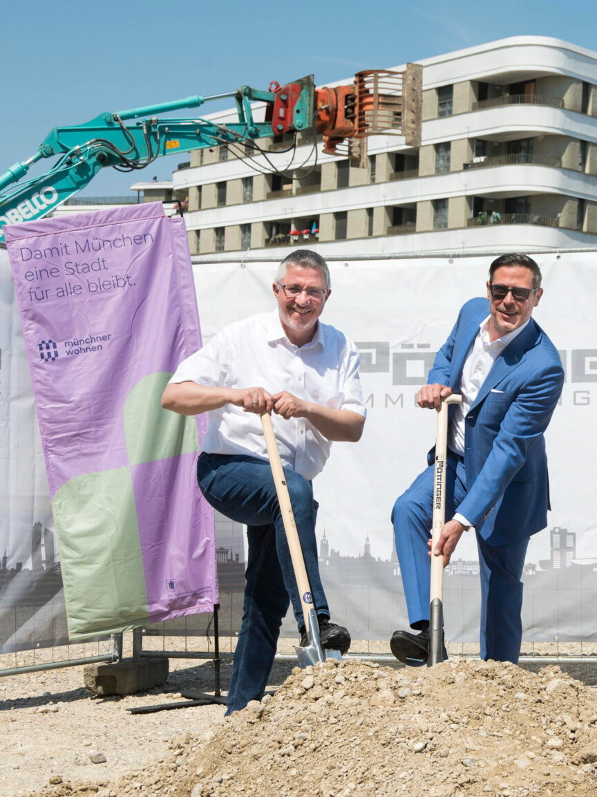 Zwei Männer setzen je einen Spaten in einen Geröllhaufen. Sie stehen auf einer Baustelle und sind umringt von Baggern und weiteren Baugeräten.