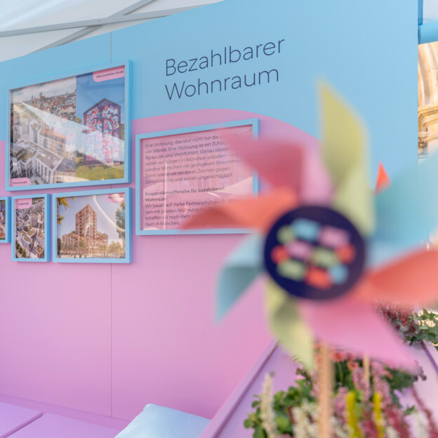 Blick auf einen Messestand auf dessen Rückwand steht "Bezahlbarer Wohnraum". Daneben hängen Fotografien von Neubauten und Quarteieren. IM Vordergrund sieht man ein Windrad mit dem Münchner Wohnen Logo.