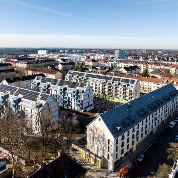 Drohnenaufnahme von oben der Wohnanlage an der Karlingerstraße. Drei Riegel stehen hintereinander und werden auf der rechten Seite von einem vierten Riegel abgeschlossen. Auf den Dächern sind Photovoltaikanlagen. Drum herum sieht man die umliegenden Wohngebäude.