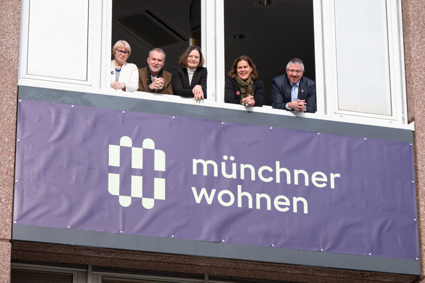 Geschäftsführung und Bürgermeisterin Verena Dietl blicken aus einem Fenster. Unter ihnen sieht man das neue Logo der Münchner Wohnen.