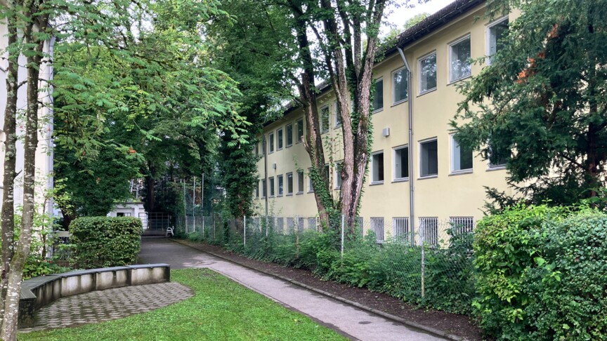 Blick auf ein dreistöckiges älteres Wohngebäude davor ein asphaltierter Weg und eine Rasenfläche.