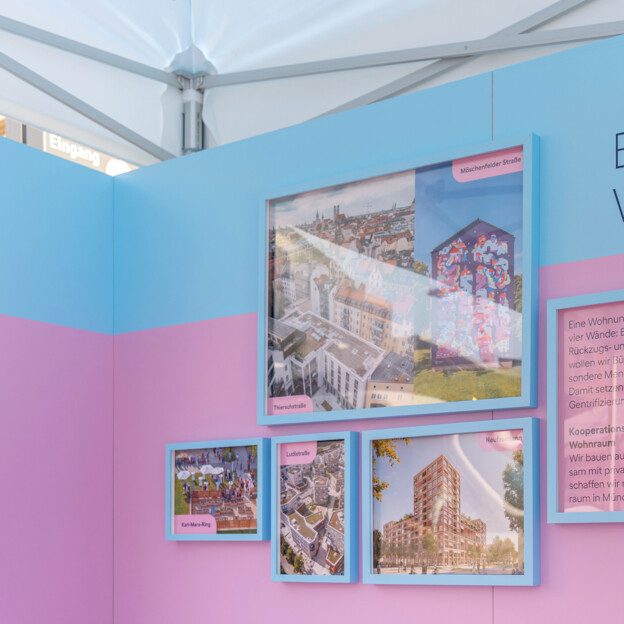 Blick auf einen Messestand auf dessen Rückwand steht "Bezahlbarer Wohnraum". Daneben hängen Fotografien von Neubauten und Quarteieren. IM Vordergrund sieht man ein Windrad mit dem Münchner Wohnen Logo.