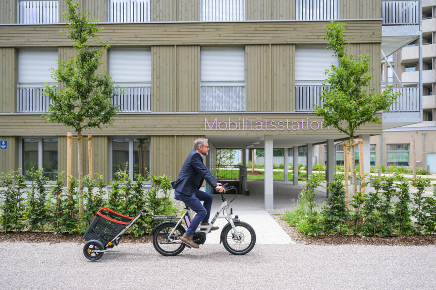 Ein Mann fährt auf einem Fahrrad mit Anhänger. Im Hintergrund sieht man ein Wohngebäude mit einer Mobilitätsstation.
