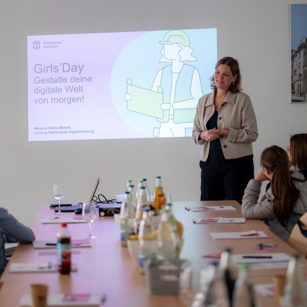 Blick über den Tisch hinweg auf eine Präsentation auf der steht "Girls' Day". Daneben steht eine Frau, um den Tisch sitzen einige junge Mädchen.