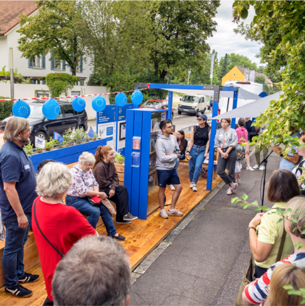 Blick auf eine Straße an der Parkplätze mit einem Holzboden überbaut sind. AUf dem Holzboden sind Sitzgelegenheiten montiert. Darauf sitzen mehrere Menschen, es hängen Luftballons über den Besuchern und jemand hält eine Ansprache.