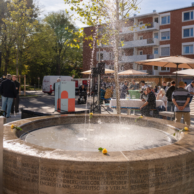 Blick auf einen Brunnen im Innenhof, der eine große Fontäne sprüht. Dahinter das Rednerpult im Münchner Wohnen Design und ein Wohnhaus.