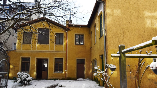 Ansicht des denkmalgeschützten Hauses, dass im Innenhof einer Wohnanlage steht. Das L-förmige Gebäudeist zweistöckig und gelb gestrichen. Die Farbe ist sieht schon älter aus und man erkennt, dass das Gebäude schon älteren Baujahres ist.