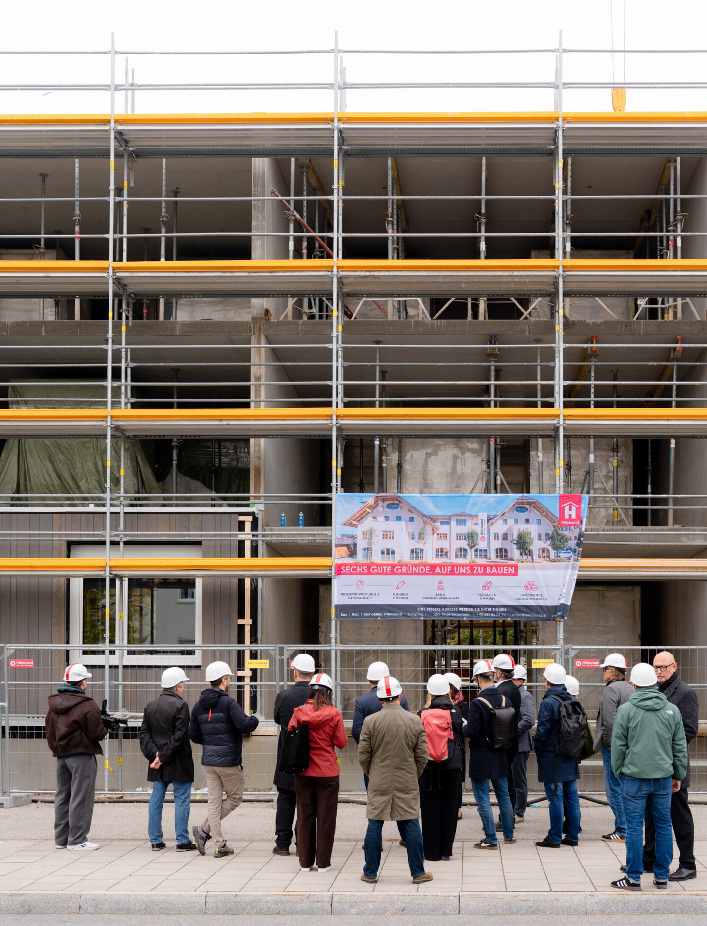 Eine Gruppe von ca. 20 Personen mit weißen Bauhelmen steht vor dem Baustellengebäude. Auf dem Plakat steht "Sechs gute Gründe, auf uns zu bauen".