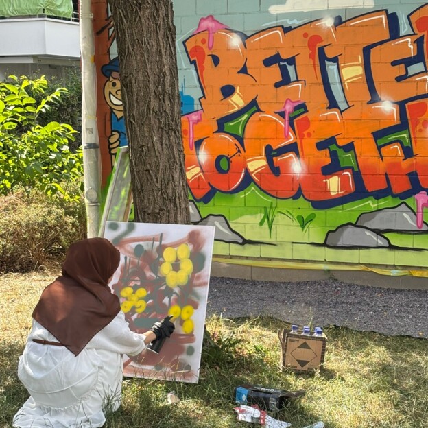 Mädchen kniet vor einer Leinwand und sprüht. Dahinter sieht man die Rückwand eines kleinen Häuschens auf der "Better together" aufgesprüht wurde.