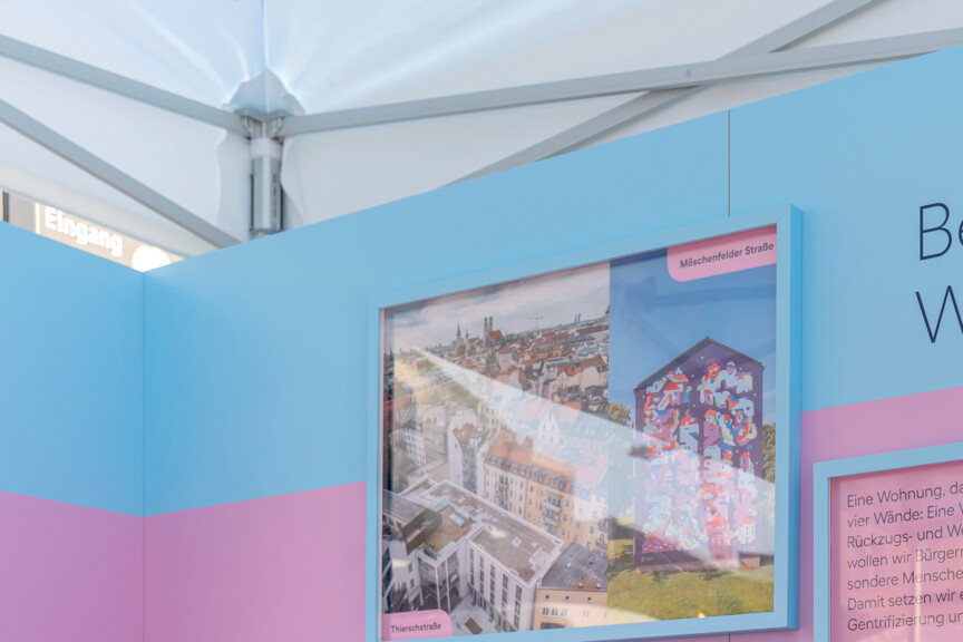 Blick auf einen Messestand auf dessen Rückwand steht "Bezahlbarer Wohnraum". Daneben hängen Fotografien von Neubauten und Quarteieren. IM Vordergrund sieht man ein Windrad mit dem Münchner Wohnen Logo.