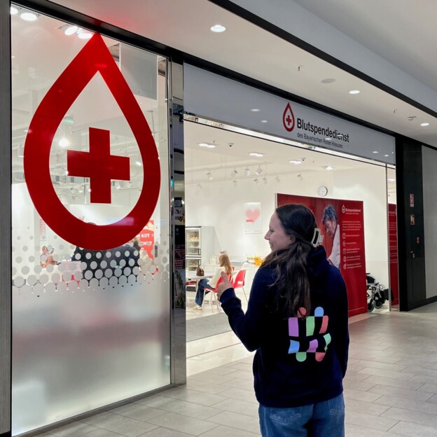 Eine Frau steht vor dem Eingang des Blutspendedienstes. Auf ihrem Pullover ist auf dem Rücken groß das Logo der Münchner Wohnen.