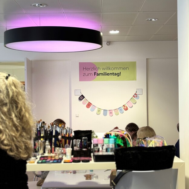 Blick auf ein Plakat auf dem "Herzlich willkommen zum Familientag" steht. Davor sieht man Schminkkoffer, Spielsachen und Kinder.