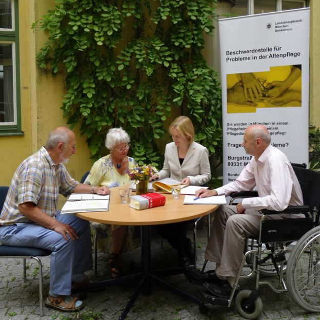 An einem Tisch im innenhof sitzen ein älteres heepaar, ein Mann im Rollstuhl und eine Frau. Hinter Ihnen steht ein Banner mit der Aufschrift "Beschwerdestelle für Probleme in der Altenpflege". Das Bild zeigt eine Beratungssituation.