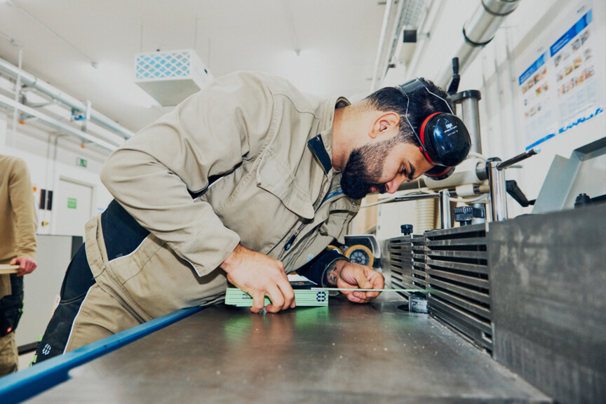 Ein Mann ist über eine Werkbank gebeugt und nimmt Maß. Er trägt Schutzkopfhörer und Arbeitskleidung.