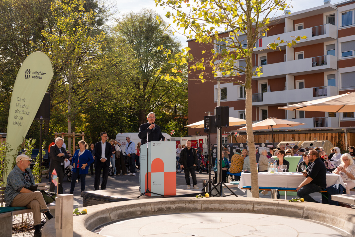 Blick auf einen Brunnen in einer Wohnanlage. Dahinter steht ein Mann am Rednerpult. Drum herum viele Menschen.