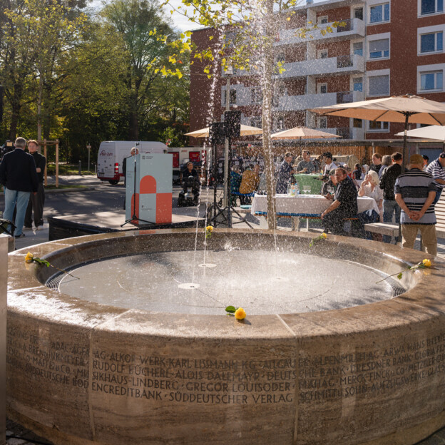 Blick auf einen Brunnen im Innenhof, der eine große Fontäne sprüht. Dahinter das Rednerpult im Münchner Wohnen Design und ein Wohnhaus.