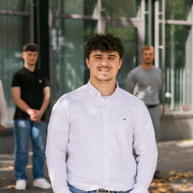 Im Vordergrund steht ein junger Mann mit dunklen Haaren. ER lächelt und trägt ein weißes Hemd. IM Hintergrund erkennt man weitere junge Menschen.