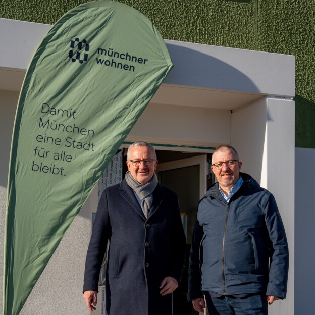Zwei Männer stehen vor einem Hauseingang. Links von Ihnen steht ein Banner der Münchner Wohnen.