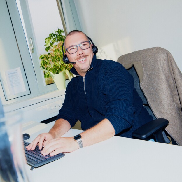 EIn Mann mit Brille und Headset sitzt am Schreibtisch und lacht in die Kamera.