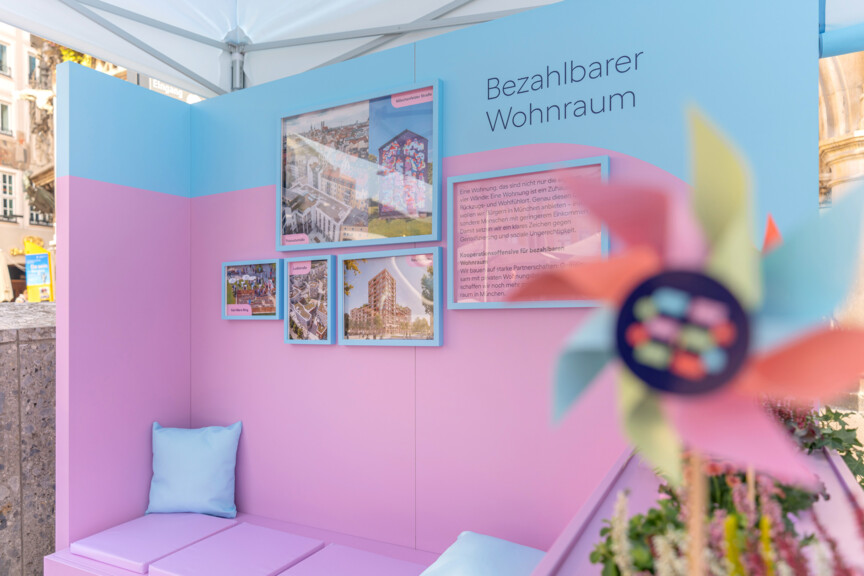 Blick auf einen Messestand auf dessen Rückwand steht "Bezahlbarer Wohnraum". Daneben hängen Fotografien von Neubauten und Quarteieren. IM Vordergrund sieht man ein Windrad mit dem Münchner Wohnen Logo.