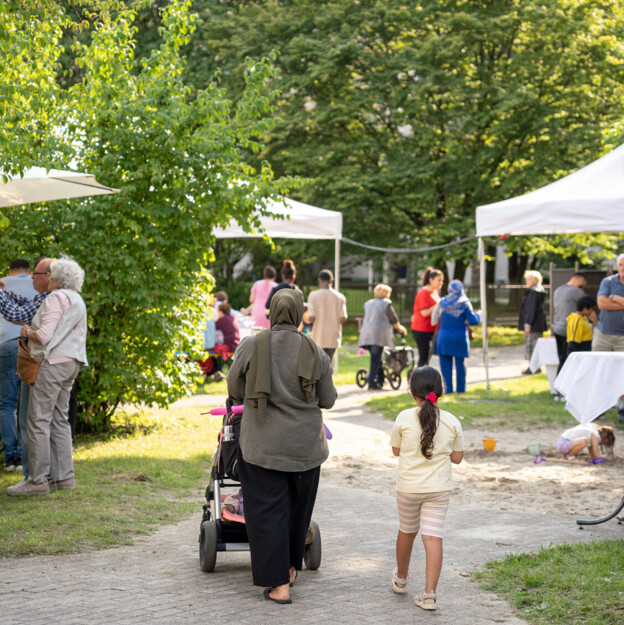 Blick in den Innenhof in dem mehrere Menschen zusammenstehen und feiern. Im Zentrum des Bildes sieht man eine Mutter mit Kinderwagen und ihrer Tochter von hinten.