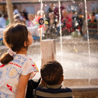 Zwei Kinder stehen am Brunnen, der kleine Fontänen sprüht. Sie pusten ein Windrad im Münchner Wohnen Logo an.