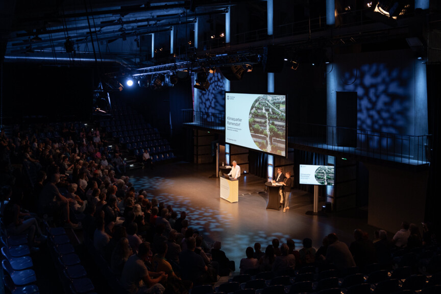 Blickpber Publikum hinweg auf den Geschäftsführer und zwei Mitarbeiter der Münchner Wohnen, die eine Präsentation zum Klimaquartier Ramersdorf halten.