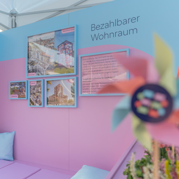 Blick auf einen Messestand auf dessen Rückwand steht "Bezahlbarer Wohnraum". Daneben hängen Fotografien von Neubauten und Quarteieren. IM Vordergrund sieht man ein Windrad mit dem Münchner Wohnen Logo.